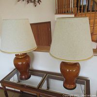 Pair of brown ceramic table lamps placed side by side on a glass top wooden table showing overall front view with beige textured fabric shades.