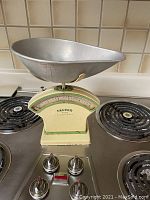 Full view of vintage Salter kitchen scale showing cream body, green trim, and large aluminum scoop on top. Positioned on a stove with four burner knobs visible.