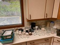 Wide view of countertop with a collection of coffee mugs, clear glass mugs, and other glassware including a basket with additional items.