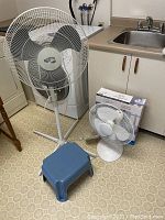 Full lot: large Hampton Bay pedestal fan, smaller Mainstays oscillating table fan with box, and small blue plastic step stool on kitchen floor near sink.
