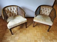 Pair of vintage cane-backed tub chairs viewed from front and slightly above, showing cane lattice backs, button tufted velvet upholstery, wooden legs with carved details on parquet floor.