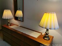 Full view of both brass table lamps placed on a wooden dresser with one lamp turned on showing the warm light and pleated white shades.