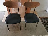 Front view of the pair of metal chairs with wooden backrests and black vinyl seats on metal legs.