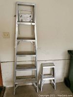Full view of silver aluminum 6-foot stepladder next to smaller two-step stool against a wall in garage with visible wear and dust.