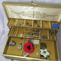 Jewelry box open to show content including rings, pins, cufflinks, keys, padlock, ceramic bottles, red flower pin, spoon and decorative heart pendant.