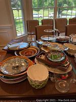 Wide view of table holding various ceramic serving platters, tiered servers, and glass plates, showing the volume and assortment of items to be included in lot.