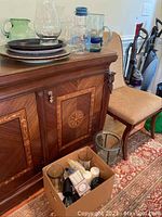 Wood cabinet topped with various glass pitchers and vases, large gold-trimmed plates, a chair, and a box with glass items and candles.