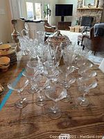 Photo showing multiple cut crystal wine and martini glasses arranged on a wooden table, with some plates stacked behind.