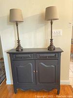 Front view of black wall table with two doors and two drawers, with two decorative lamps on top.