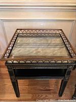 Front view of the square end table showing the glass top with metal circular design frame, black wood structure and gold decorative accents.