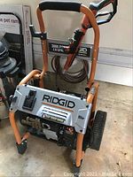 Front view showing orange frame, silver panel with Ridgid logo, mounted high-pressure hose
