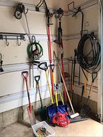 Photo showing various garden tools leaning against a garage wall including shovels, rakes, brooms, and a garden hose hung on the wall.