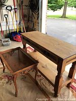 Both rustic wooden foyer table and carved end table with glass insert shown in a garage setting on a floral rug.