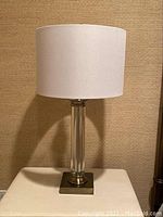 Full view of a table lamp with white drum shade, clear glass column, and brass metal base.