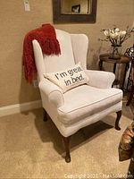 Full view of cream upholstered wingback chair with a red throw blanket on the back and a cushion on the seat.