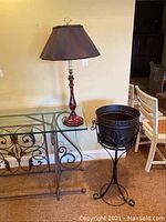 Table lamp and black metal plant stand with removable planter on the floor against a wall.