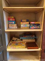 Image shows multiple boxed games arranged on wooden shelves including Sports Challenge for Kids, Smart Ass, Fact or Crap, The Game of Life, Battle of the Sexes, Where in the World?, Great States, Monopoly, Taboo, Word Whiz, and a checkers set.
