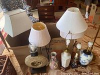 Front view of four table lamps, one floor lamp, three shades, and one decorative vase arranged side by side on carpet.