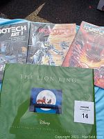 Photo showing three Robotech books and green folder for The Lion King lithographs on blue cloth.