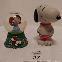 Front view of Peanuts snowglobe with Lucy inside globe holding football and Snoopy and Woodstock figures on green base, next to plastic Snoopy coin bank.