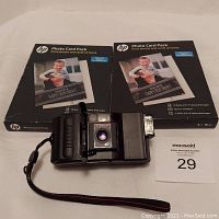 Photo showing Chinon 35 mm camera with two HP Photo Card Packs behind it, all items displayed on a white surface with auction lot number card visible.