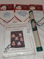 Photo of three sets of holiday cross stitch napkins (Santa, Rose, Poinsettia), sealed roll of Aida cloth, and Laura Doyle Designs Santa Ornament kit with packaging visible.