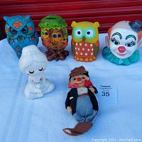 Front view of all six items on display against red backdrop showing owl banks, Yoga Girl bank, clown bank and Hobo doll