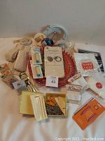 Photo of overall lot with various knitting, cross stitch, crochet, and quilting supplies. Items include pink Knit Quick knitting loom, spools of silk thread, lace trims, needles, pins, scissors, and packaging materials.