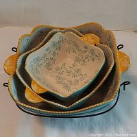 Top angled photo showing three floral lace patterned ceramic bowls in teal and yellow colors nested inside each other in a black wire rack.