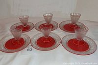 Set of six etched glass plates with matching footed cordial glasses arranged together, each plate with a well holding a glass, all on white surface.