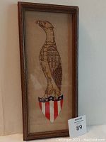 Framed vertical cross stitch of an eagle on a patriotic shield. The eagle is brown with detailed stitching, perched on a shield with red and white stripes and a blue top with white stars. The frame is wood with visible grain and the fabric shows some water staining lower left.