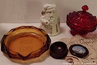 Fenton ruby red hobnail footed covered dish, Santa candle cover, Viking Glass amber ashtray, plastic box with sailing ship lid, and doilies all arranged on surface.
