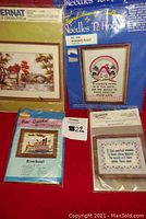 Photo showing fronts of four needlework kits with illustrations: 'Cider Mill', 'Woman's Place', 'Riverboat', and 'Clean Houses' pattern titles visible.