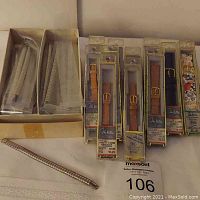 Overview of 12 stainless steel Bulova stretch watchbands in original packaging and loose stretched band, with 8 John Weitz leather bands in original packaging in background
