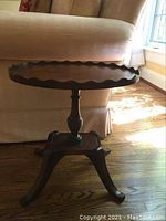 Side view of the antique wooden side table showing the undulating pie crust edge, turned pedestal, and four curved legs.