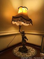 Full view of the vintage brass cherub table lamp lit, showing the fabric shade with fringe and the cherub base on a wooden corner table.