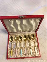 Six gold-tone spoons with polished bowls and colored handles arranged in red box lined with cream satin fabric, showing front view with handles pastel yellow to white.