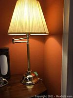 Full view of lit night table lamp showing brass base, tall column, swivel extendable arm and beige pleated fabric shade.