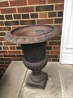 Front side view of cast iron planter urn showing decorative rim and pedestal base against brick and concrete background.