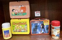 Photo showing four lunch pails and two thermos bottles on a wooden shelf.