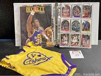 Binder with a vintage Beckett Basketball Monthly magazine and pages filled with basketball trading cards, along with a yellow Lakers onesie laid in front.