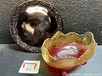 Two vintage colored glass serving items: a dark red round glass plate with ornate leaf and bead border, and an orange-red floral textured footed glass bowl with scalloped edge.