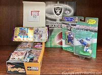 Box containing multiple unopened Upper Deck Collector's Choice football card packs with visible cards in plastic wrapping, an Oakland Raiders poster behind the box, and a Starting Lineup figurine of a player with jersey number 50 still in original packaging.