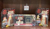 Two Star Wars Episode I action figures in packaging with Clash of the Lightsabers card game box between them on shelf.