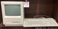 Photo showing front view of Apple Macintosh SE computer with built-in monitor, connected Apple wired keyboard, and case or stand underneath them on wooden shelf.