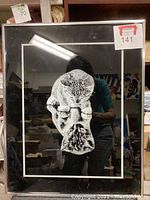 Front view of the framed print showing a cat looking at its reflection on a black background, with thin metal frame and reflection of room behind glass.
