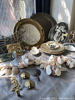 Photo of mixed mermaid and seashell decorative items displayed on table including brass mirrors, etched mirrors, shell garland, porcelain plaque, brass hooks, butterfly wall hangings, and embroidered patch.