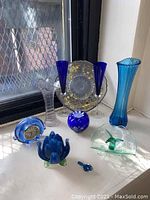 Mixed blue glass collectibles including vases, figurines, plates and flutes arranged on a windowsill
