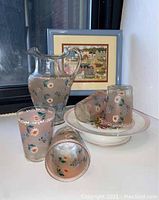 Clear glass pitcher and four matching glasses, two glasses on a floral tray, framed print behind