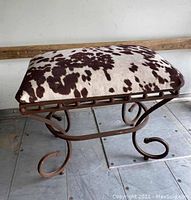 Front side view of bench showing metal scroll legs and cowhide patterned cushion.